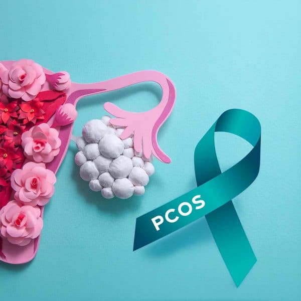 A teal ribbon labeled PCOS is next to an artistic representation of ovaries, with one ovary decorated with white cotton balls and pink flowers, symbolizing women’s health and polycystic ovary syndrome awareness.