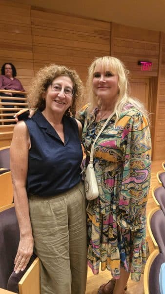 Two women smile for a photo in an auditorium. One wears a navy sleeveless top and beige pants; the other has a colorful patterned dress and a white purse. Wooden walls and purple seats are visible.