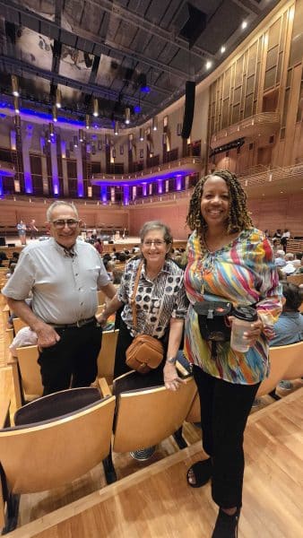 Three people stand and smile inside a modern concert hall with wooden seats and blue lights on stage. They are dressed casually; one holds a drink cup. Other audience members are seated behind them.
