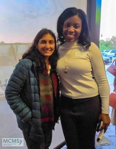 Two women smiling and standing together indoors, one wearing a jacket and scarf, the other in a white top. There is a logo in the bottom left corner that reads MCMS..