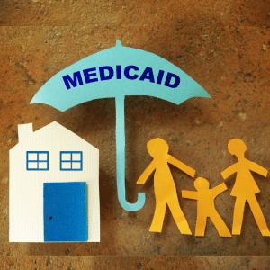Paper cutouts of a house and a family under a blue umbrella labeled MEDICAID on a brown background, symbolizing health coverage and protection.