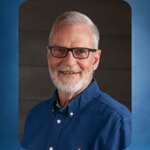 Smiling older man with short gray hair, beard, and glasses, wearing a blue button-up shirt. He is posed in front of a dark background with rounded corners and a blue frame.