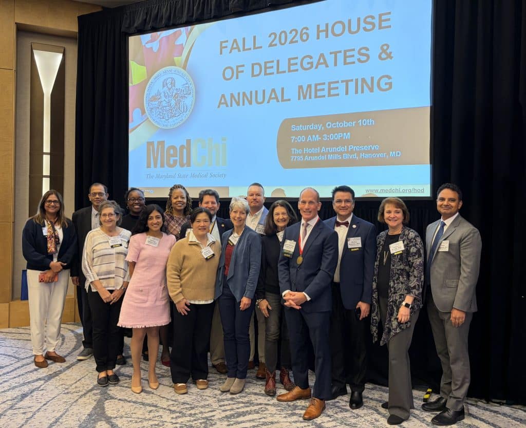 A group of fourteen people stand in front of a projection screen announcing the Fall 2026 MedChi House of Delegates and Annual Meeting at The Hotel Arundel Preserve.